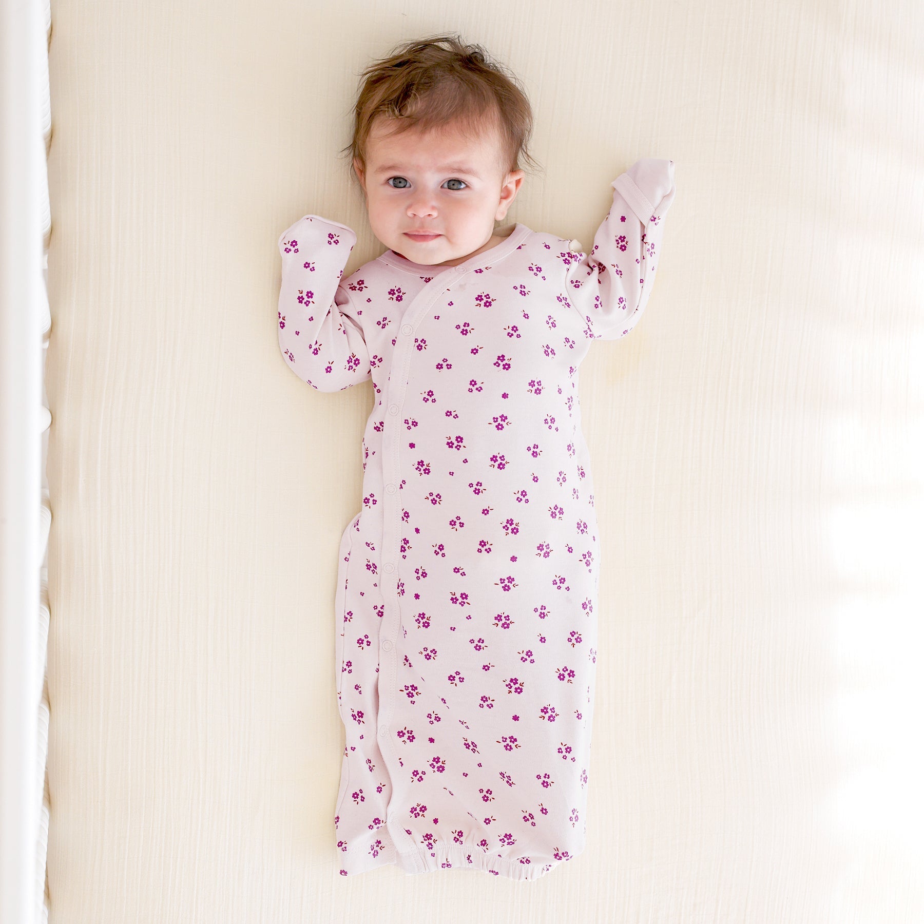 Baby wearing a light purple sleep gown with purple floral patterns lying on a white crib.